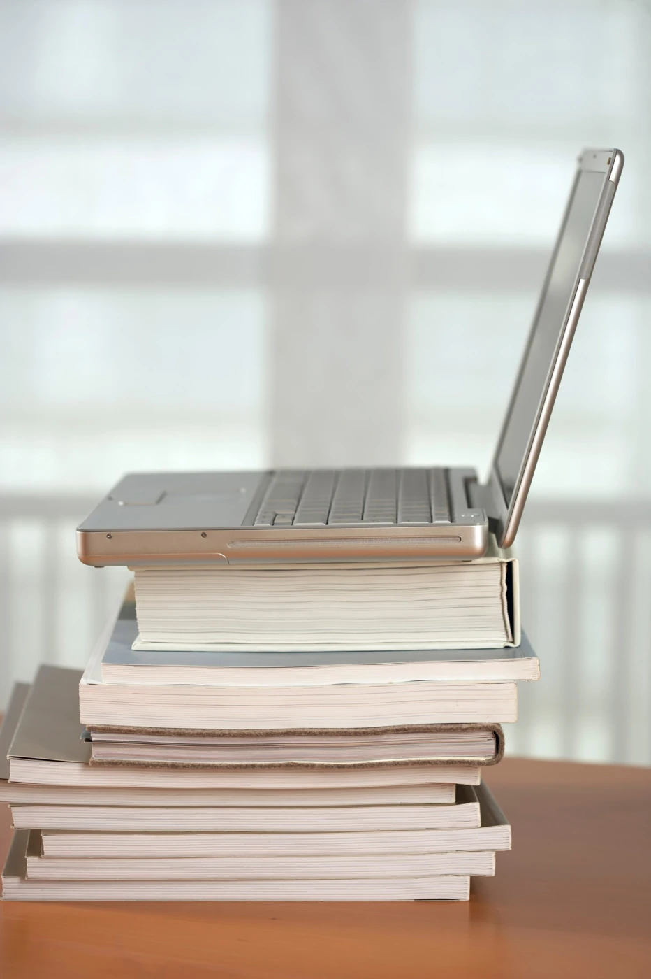 An open laptop rests on a stack of books beside a window.