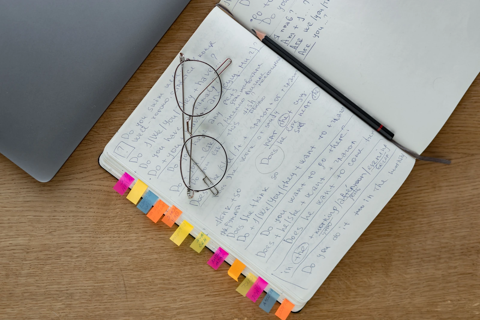 Eyeglasses and a pencil rest on a notebook filled with handwritten notes beside a laptop.