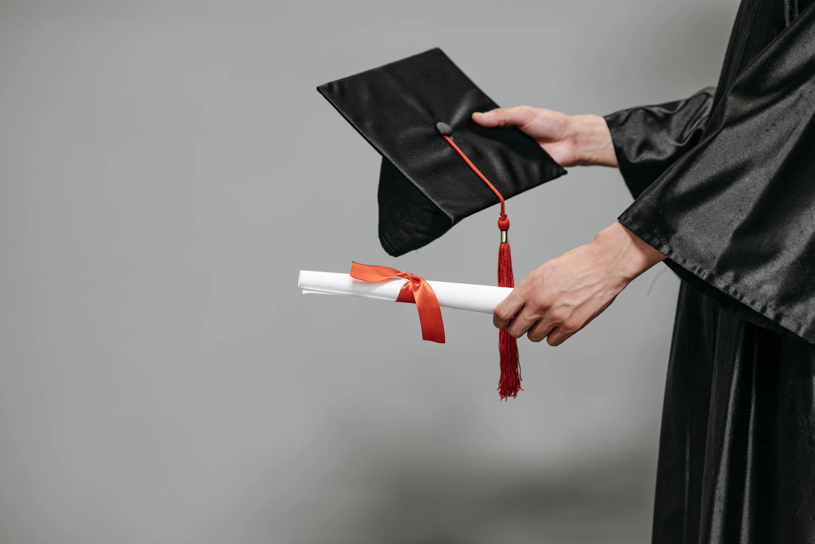 Hands in graduation robes hold a black mortarboard and a rolled diploma tied with a red ribbon.