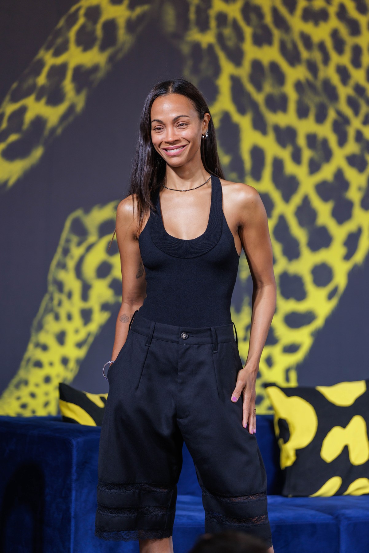 Zoë Saldaña smiles before a yellow-and-charcoal Locarno Film Festival backdrop, wearing a black sleeveless top and black shorts.